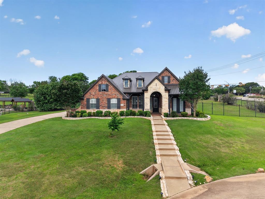 Daytime front elevation of 102 W Carruth Lane: a brick-and-stone home with a gabled metal roof and arched stone entry, set behind a stepped stone walkway up a sloping lawn