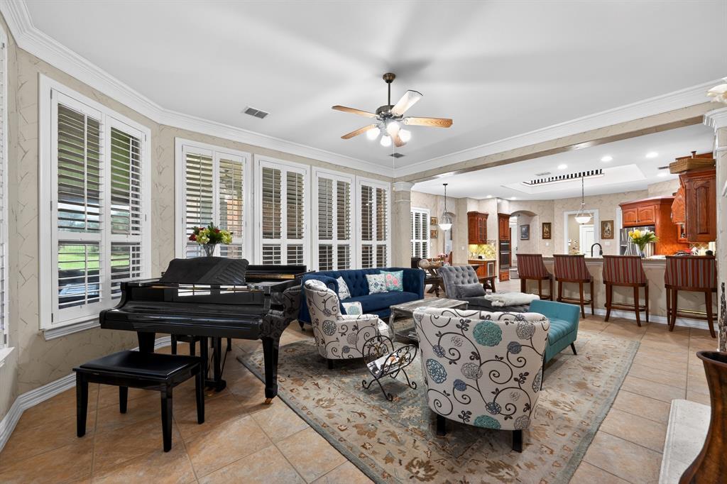 An open living area at 102 W Carruth Lane with a grand piano beside a bank of shuttered windows, flowing past columns into the kitchen and breakfast bar beyond