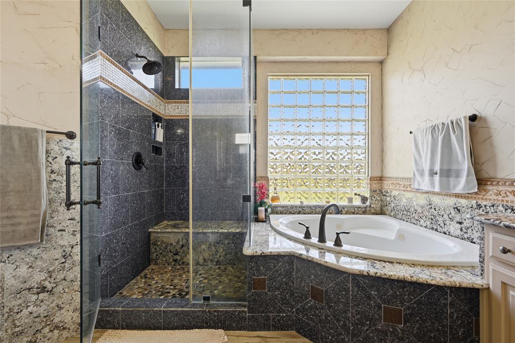 The primary bathroom at 102 W Carruth Lane with a walk-in slate-tiled shower with a bench, a corner jetted tub, granite surrounds, and a glass-block window