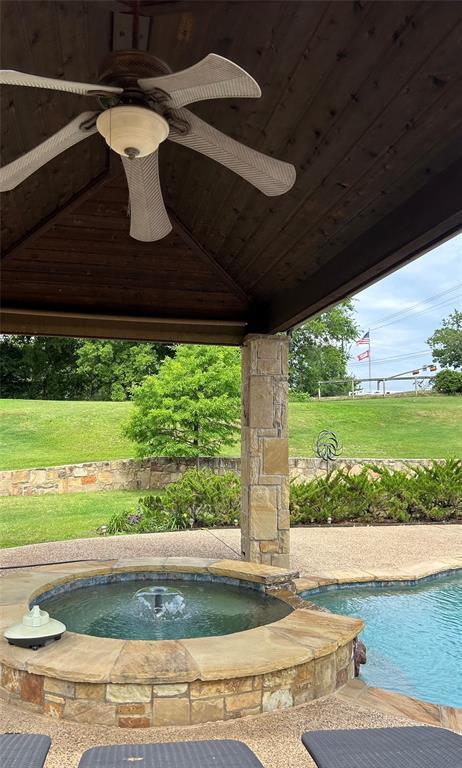 The raised stone spa at 102 W Carruth Lane with a bubbling fountain, viewed from beneath the cabana's wood ceiling and ceiling fan, looking out over the green lawn