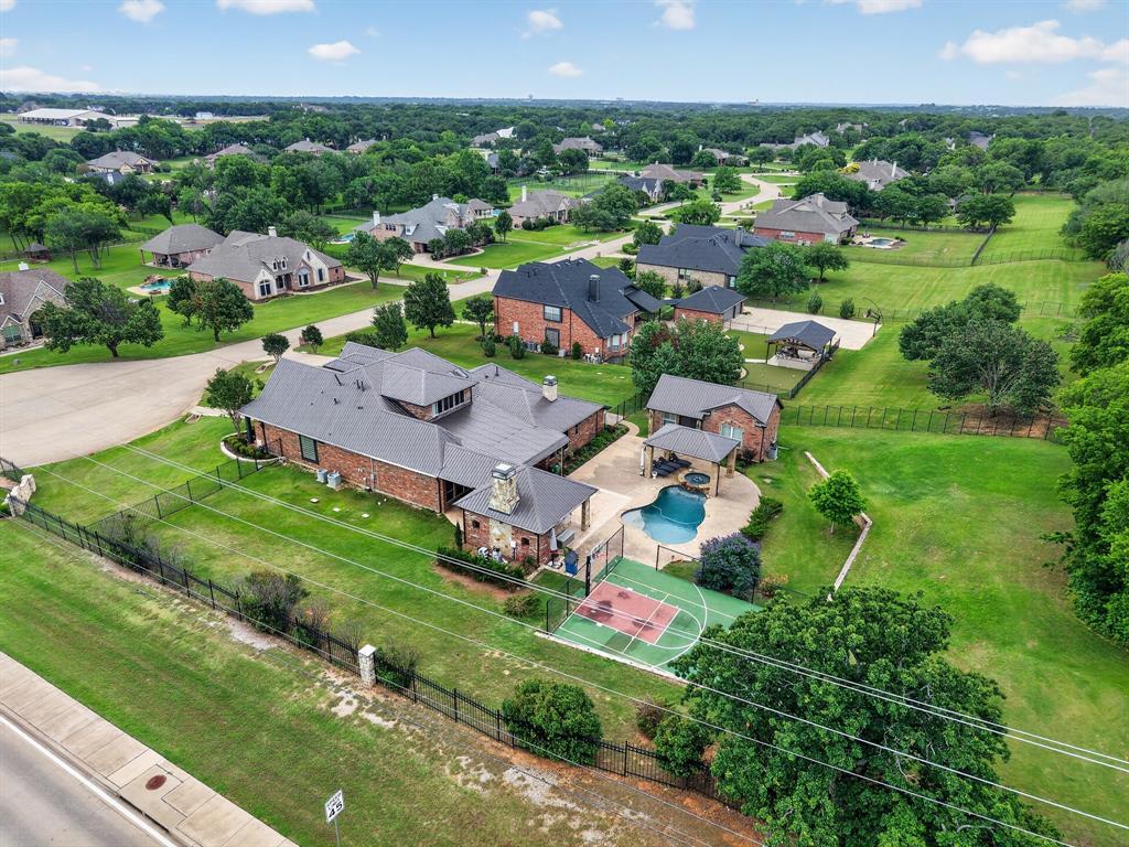 An aerial view of 102 W Carruth Lane showing the full acre: the metal-roofed home, the pool and cabana, the sport court, and lawn stretching to the wrought-iron perimeter fence