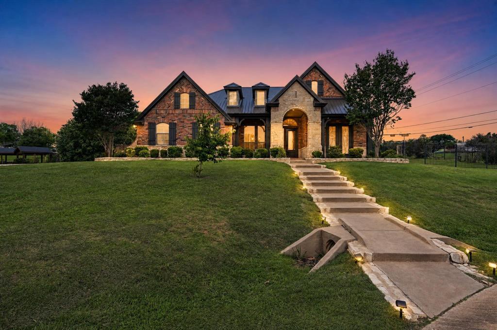 102 W Carruth Lane at dusk, a brick-and-stone Texas estate with a metal roof and lit windows above a lawn with an illuminated stone stairway, under a pink and violet sunset